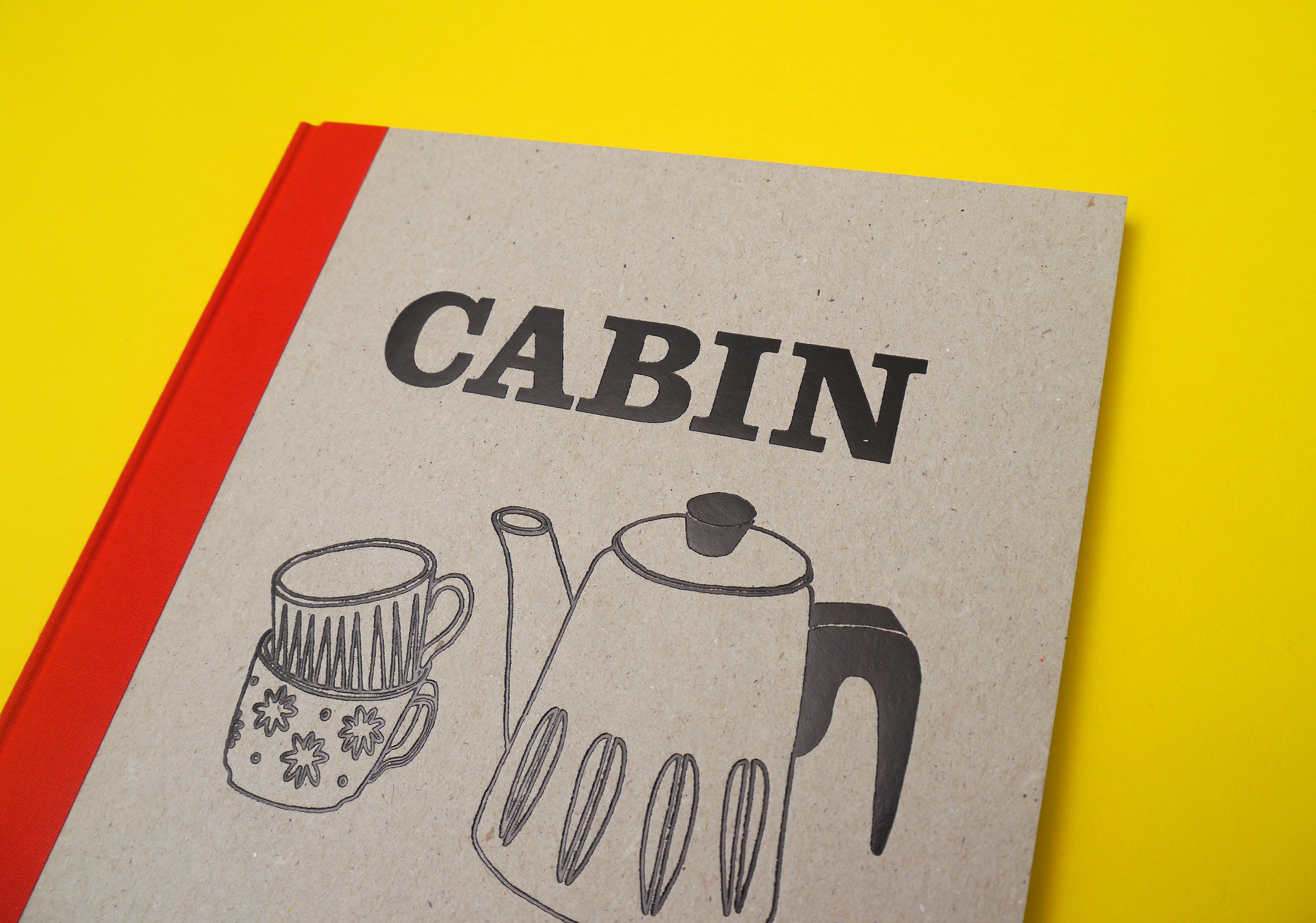 Hardback book with a cardboard cover and red spine against a yellow background, the title in black reads Cabin, with an illustration of a coffee pot and two cups